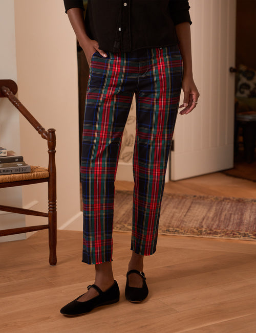 A person wearing plaid cropped pants in red, green, and navy with a black top and black flats, standing in a home interior with wood floors and a chair visible., front view