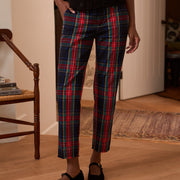 A person wearing plaid cropped pants in red, green, and navy with a black top and black flats, standing in a home interior with wood floors and a chair visible.