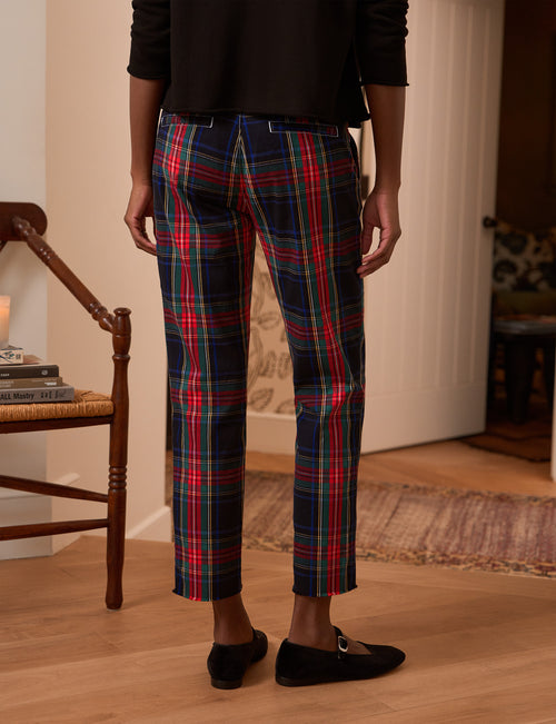 A person shown from the back wearing plaid cropped pants in navy, red, green, and yellow, paired with a black top and black flats, standing on a wooden floor in an indoor room., alternate view