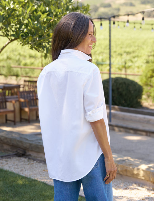 Woman wearing a white shirt and blue jeans standing outdoors with greenery in the background, alternate view