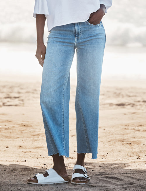 Person wearing light blue jeans and white sandals on a wooden floor., front view