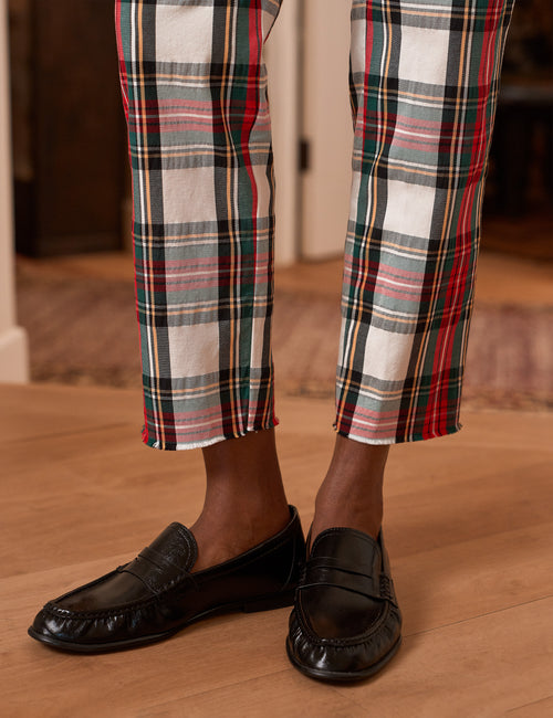 A close-up of a person's legs wearing plaid trousers with red, green, black, and white stripes, paired with black leather penny loafers on a light wood floor., view 3