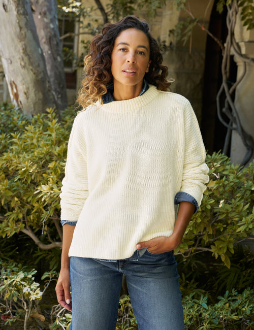 A woman wearing a cream-colored ribbed knit sweater and blue jeans, standing outdoors in front of green foliage and a tree trunk., front view