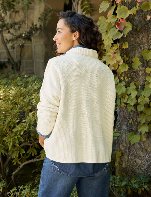 A woman wearing a cream ribbed knit mock neck sweater and blue jeans, standing outdoors next to a tree with green and red autumn leaves., alternate view