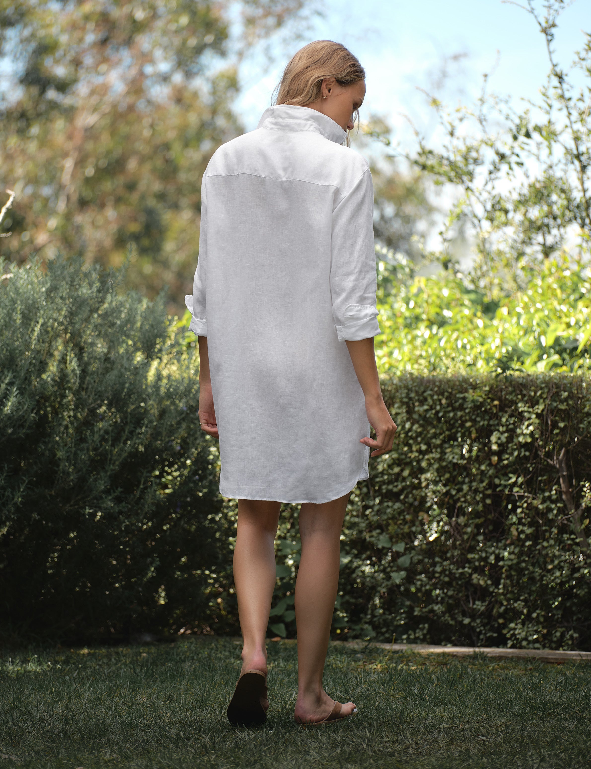 Woman wearing a white dress walking outdoors with greenery in the background, alternate view