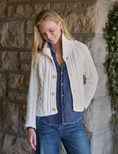A woman wearing a cream cable-knit cropped cardigan over a blue denim shirt and jeans, standing against a rustic stone wall., front view
