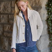 A woman wearing a cream cable-knit cropped cardigan over a blue denim shirt and jeans, standing against a rustic stone wall.