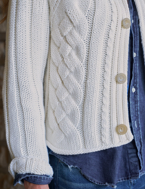 Close-up of a person wearing a chunky cream cable-knit cardigan with beige buttons, layered over a blue denim shirt and jeans., view 3