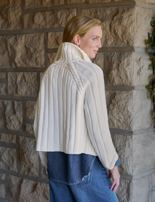 A woman wearing a cream ribbed knit sweater and blue jeans, standing in front of a stone wall., alternate view