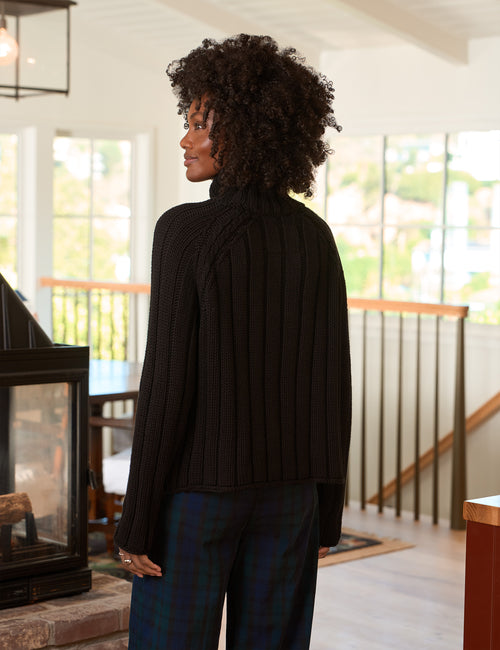 A woman with curly black hair is shown from the back wearing a black ribbed turtleneck sweater and dark plaid pants in a bright, modern home interior., alternate view