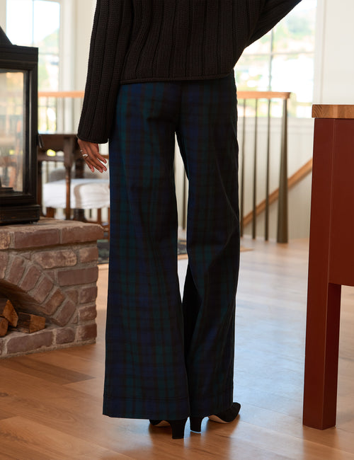 A person shown from the back wearing dark wide-leg plaid trousers and a black ribbed sweater in a home interior with wood floors and a fireplace., alternate view