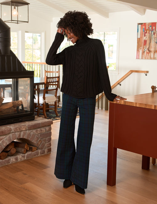 A woman stands in a bright indoor room wearing a black cable-knit turtleneck sweater and dark blue plaid wide-leg pants, photographed from the back., view 4