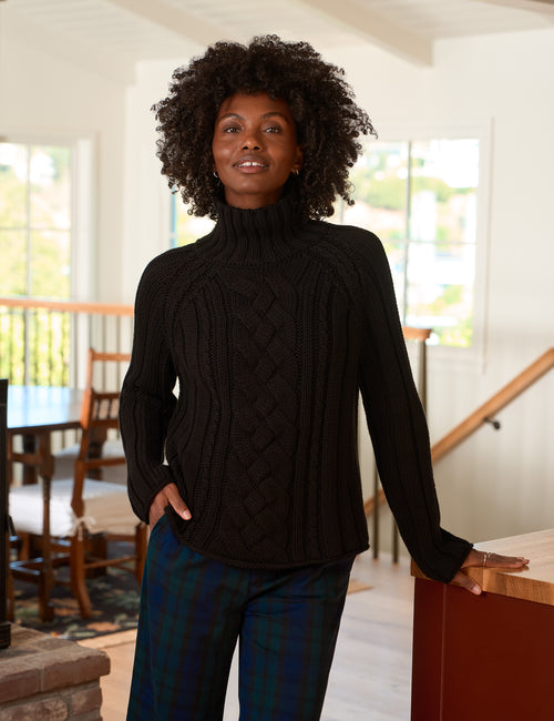 A woman with curly black hair wearing a black cable-knit turtleneck sweater and blue plaid pants, standing in a bright, modern home interior., front view