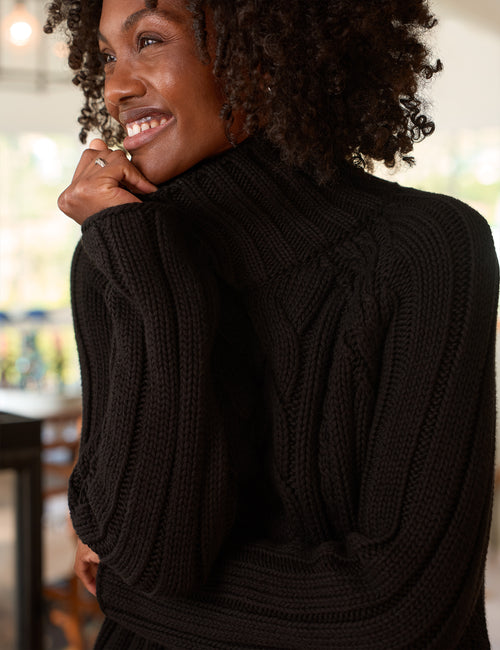 A woman with curly dark hair wearing a black ribbed knit turtleneck sweater, shown from a side-back angle in a softly lit indoor setting., view 3