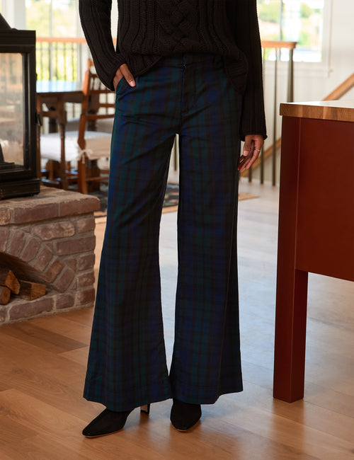 A person wearing dark plaid wide-leg pants with a black cable-knit sweater and black ankle boots, standing in a home interior with wood floors and a fireplace visible., front view