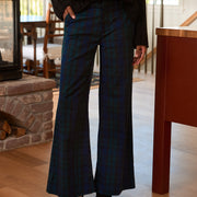 A person wearing dark plaid wide-leg pants with a black cable-knit sweater and black ankle boots, standing in a home interior with wood floors and a fireplace visible.
