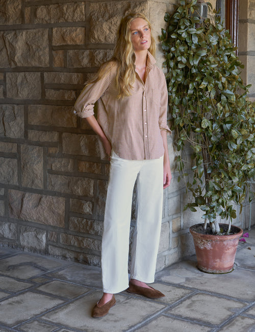 A woman stands on a stone patio wearing a beige and white gingham button-down shirt, white wide-leg pants, and brown loafers, next to a potted green plant., view 5