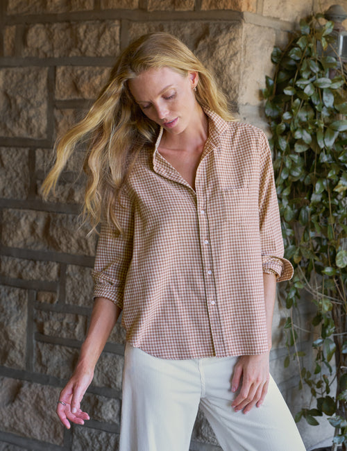 A woman wearing a brown and beige gingham button-down shirt with rolled-up sleeves and white pants, standing in front of a stone wall with green ivy., front view