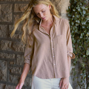 A woman wearing a brown and beige gingham button-down shirt with rolled-up sleeves and white pants, standing in front of a stone wall with green ivy.
