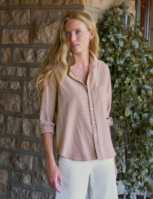 A woman wearing a light beige and white gingham button-down shirt with rolled-up sleeves, paired with off-white pants, standing in front of a stone wall and green ivy., view 4