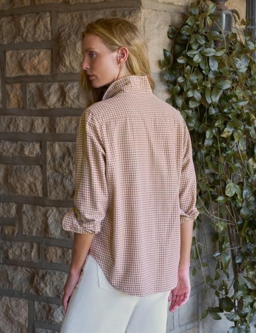 A woman wearing a light beige and white gingham check shirt with rolled-up sleeves, standing in front of a stone wall with green ivy., alternate view