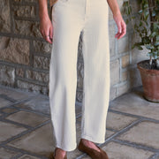 A person wearing high-waisted, barrel-leg cream trousers with a beige gingham shirt and brown suede flats, standing on a stone patio in front of a stone wall.