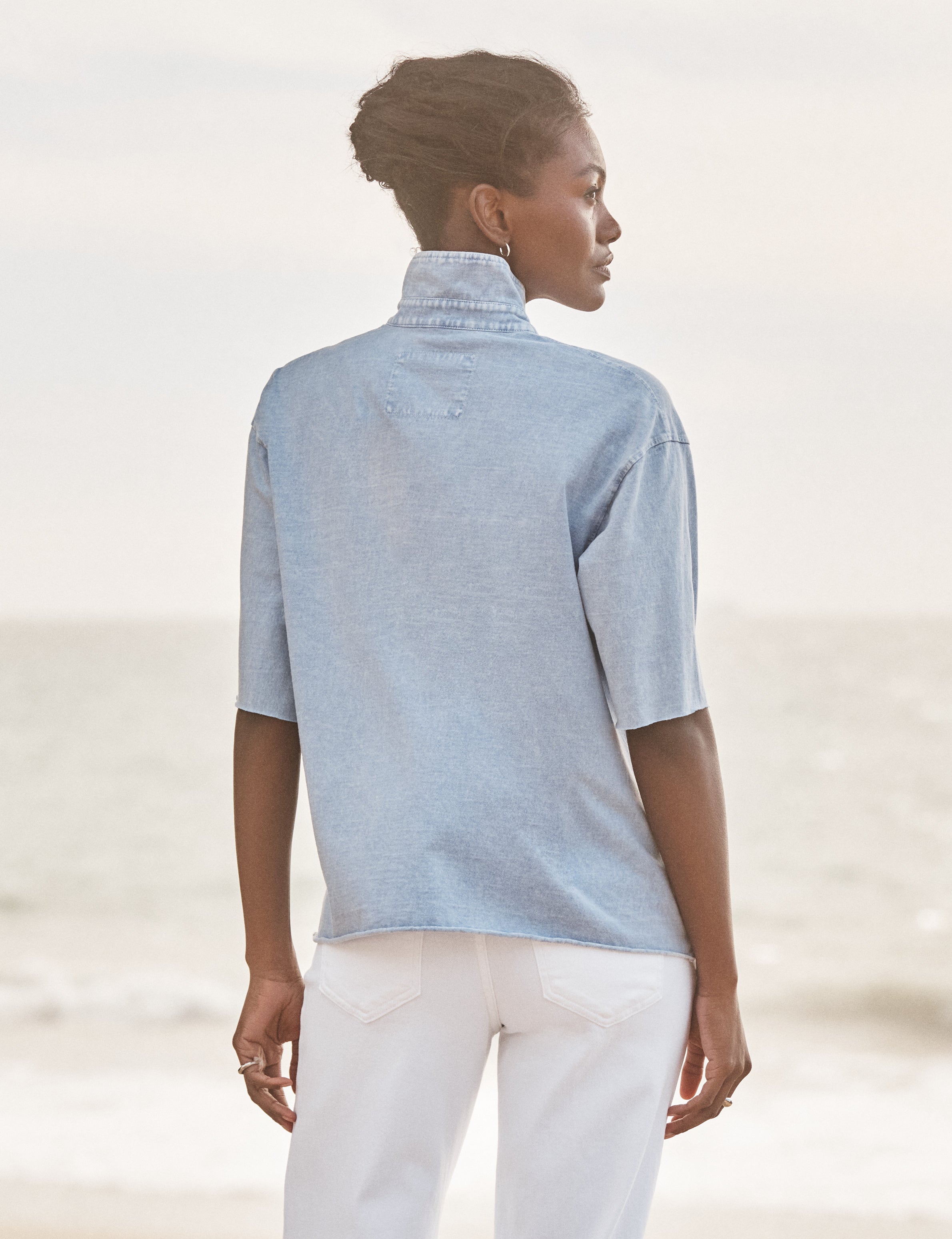Woman wearing a light blue shirt and white pants against a blurred natural background, alternate view