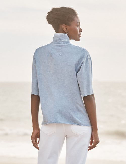 Woman wearing a light blue shirt and white pants against a blurred natural background, alternate view