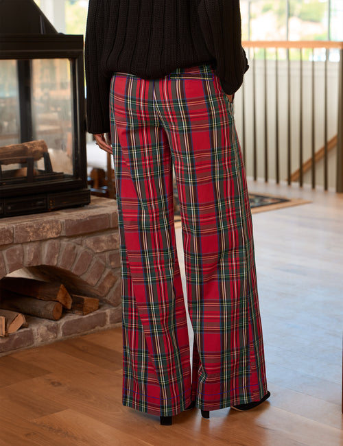 A person wearing red plaid wide-leg pants with green and yellow stripes, paired with a black ribbed sweater, standing in a home interior near a fireplace., alternate view
