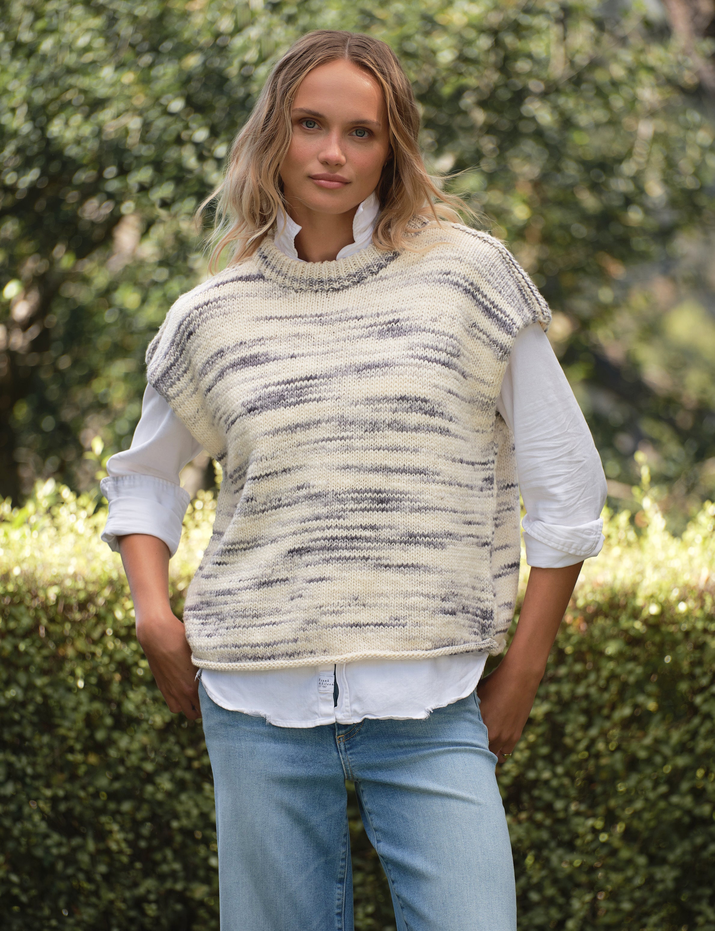 Woman wearing a patterned vest over a white shirt with blue jeans outdoors., front view