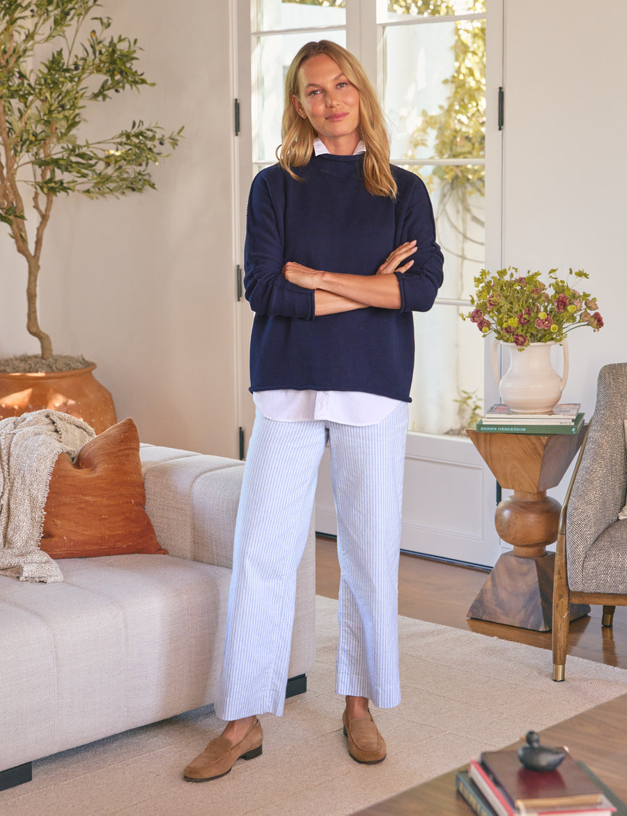 Woman standing in a living room wearing a navy sweater and white pants.