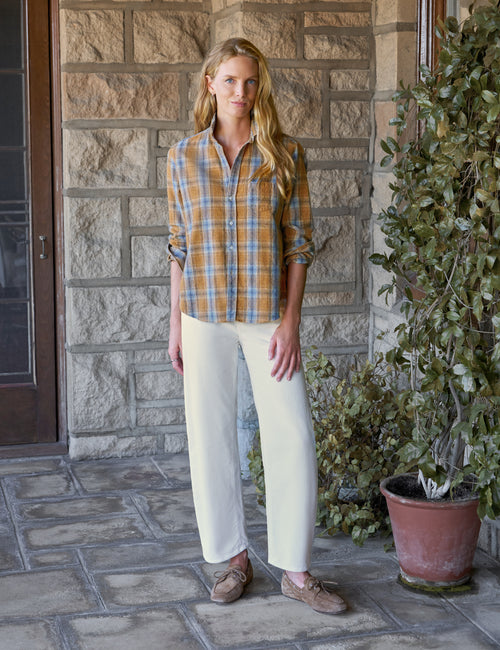 A woman stands on a stone porch wearing a yellow and blue plaid button-up shirt, cream barrel-leg pants, and brown suede loafers, with a stone wall and potted greenery in the background., view 4