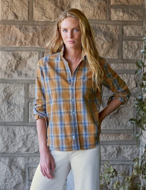 A woman stands in front of a stone wall wearing a long-sleeve plaid button-up shirt in mustard yellow, blue, and white, paired with off-white pants., front view