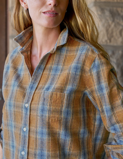 A woman wearing a long-sleeve button-up plaid shirt in brown and blue tones, photographed from the chest up against a softly blurred rustic background., view 3