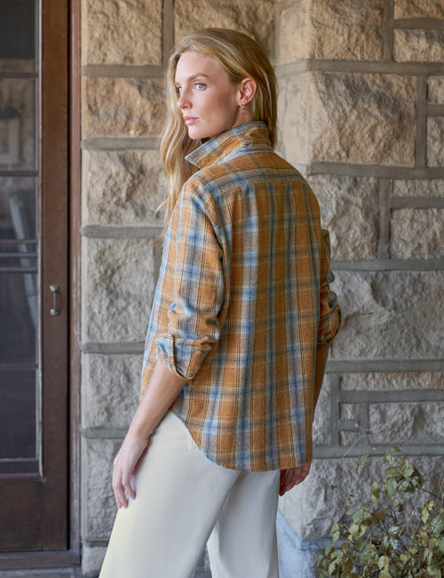 A woman wearing a long-sleeve plaid button-up shirt in mustard yellow, blue, and white, paired with cream pants, standing in front of a rustic stone wall., alternate view