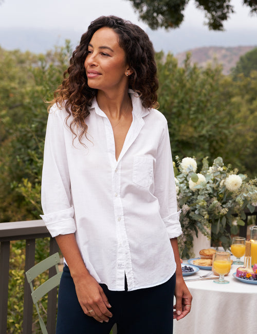 A woman wearing a white button-down shirt with rolled sleeves and a chest pocket, standing on a balcony with a garden view and a decorated table in the background., front view