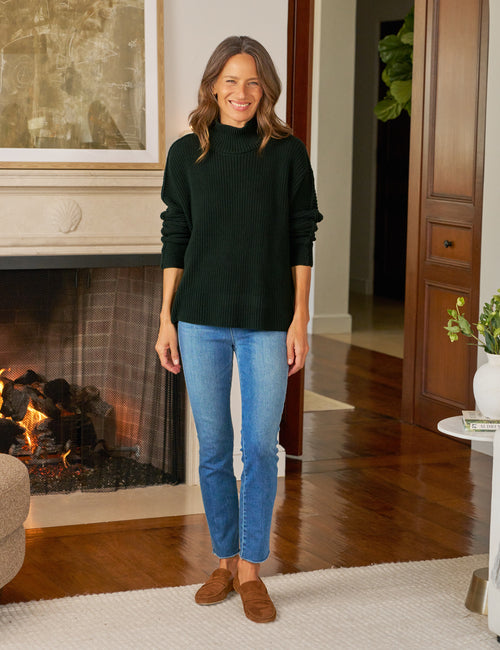 Woman wearing a dark green sweater and blue jeans standing in a living room with a fireplace., view 4