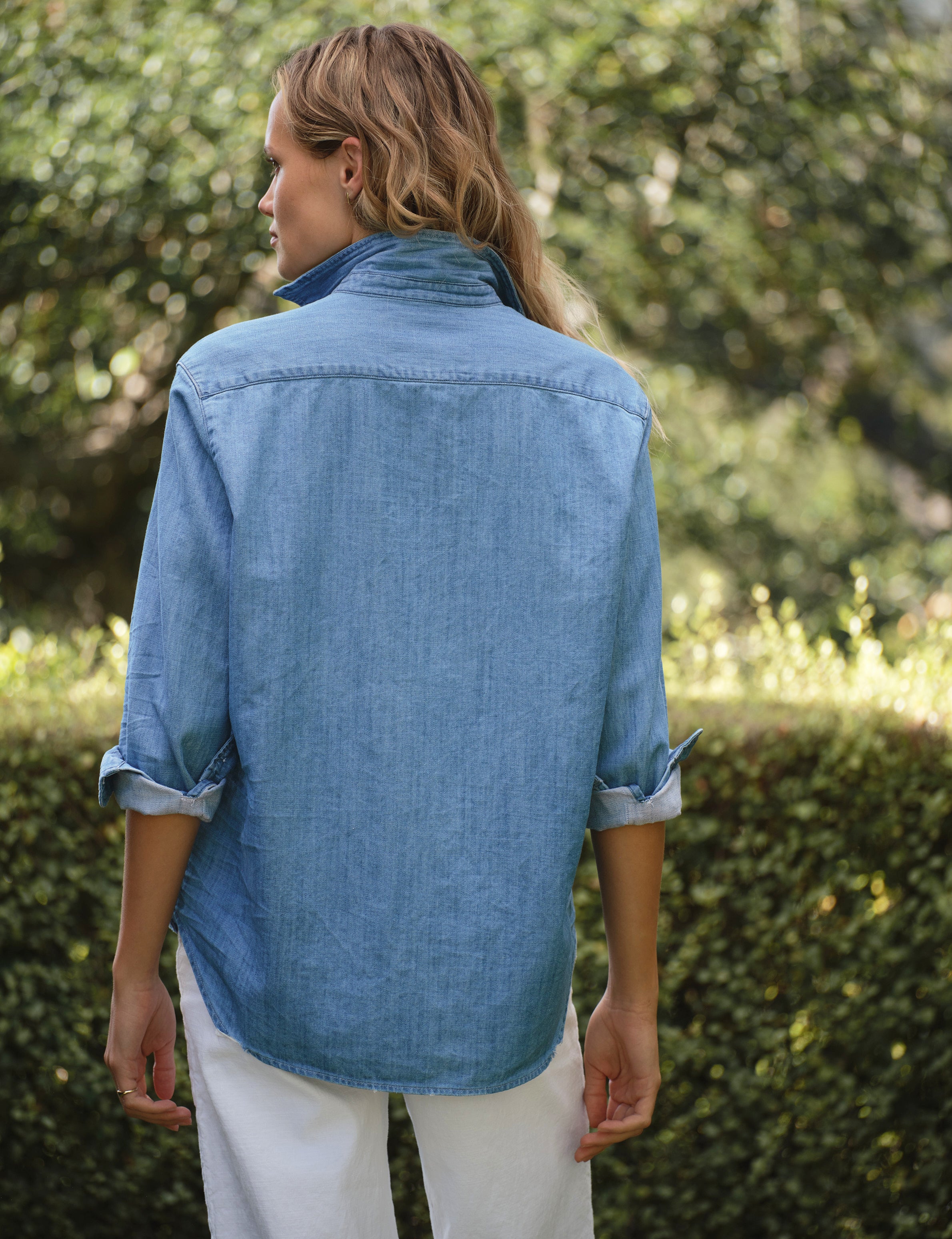 Woman wearing a blue denim shirt and white pants standing outdoors with greenery in the background, view 3