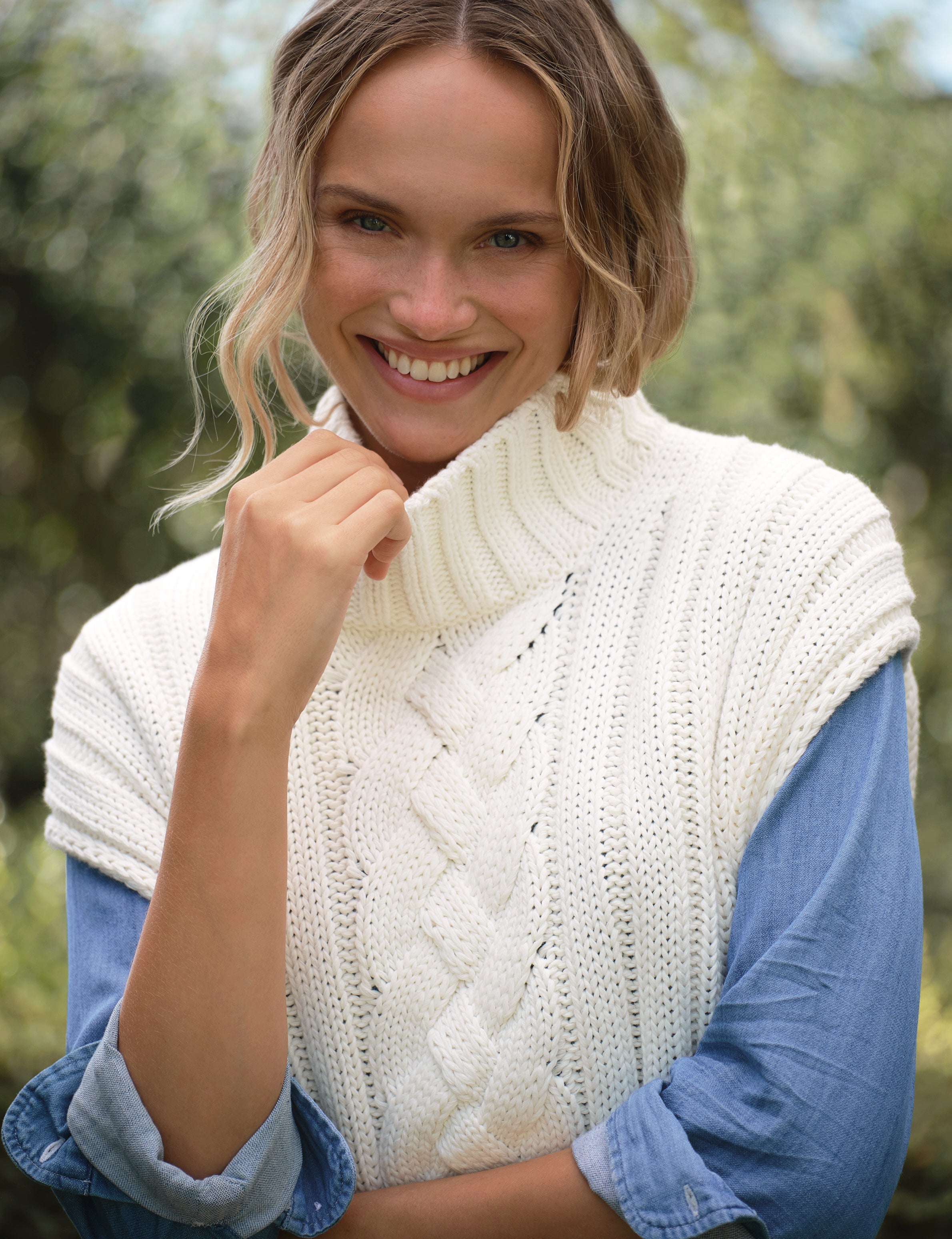 Woman wearing a white knitted vest over a blue shirt with a blurred natural background, view 4