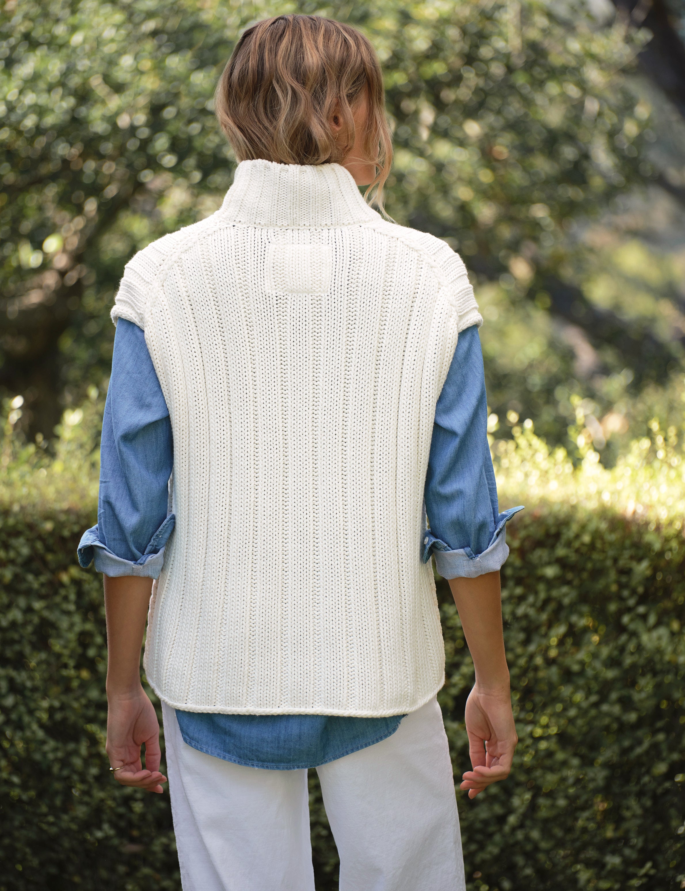 Person wearing a white knit vest over a blue shirt with a blurred natural background, view 3