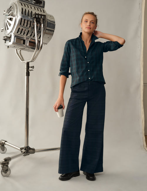 A woman modeling a dark teal plaid button-up shirt and matching wide-leg trousers, standing in a studio next to a large vintage-style light fixture., view 5