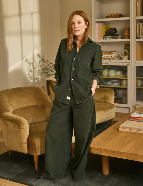 Woman in a dark green outfit standing in a cozy living room with beige armchair and wooden coffee table., front view