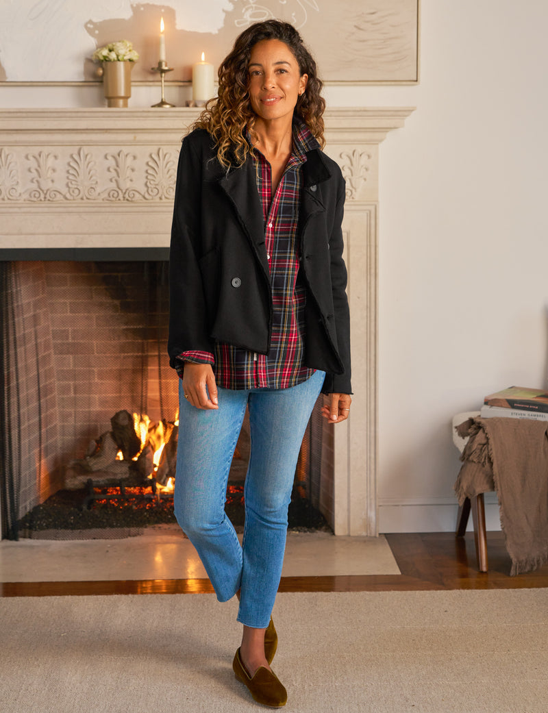 Woman standing in a cozy room with a fireplace, wearing a black coat, plaid shirt, blue jeans, and brown shoes.