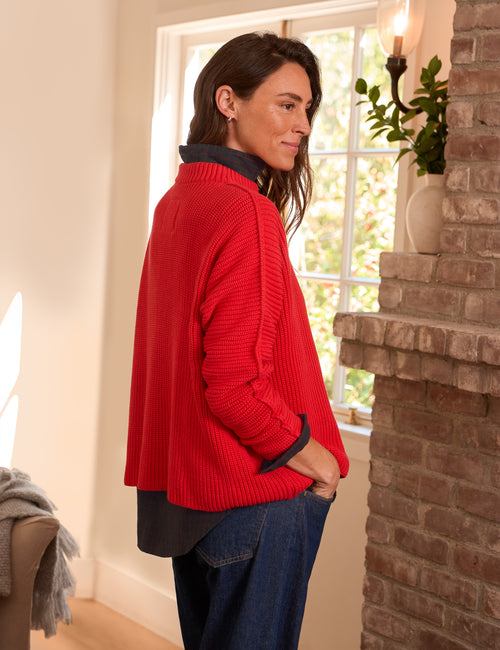 A woman wearing a bright red ribbed knit sweater, paired with blue jeans, standing in a warmly lit room next to a brick fireplace., alternate view