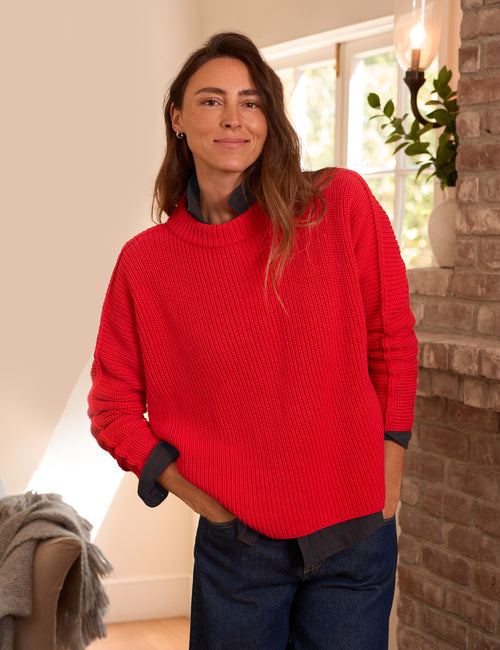A woman wearing a bright red ribbed knit sweater over a dark navy collared shirt, paired with dark blue jeans, standing in a warmly lit room with a brick pillar and window in the background., front view