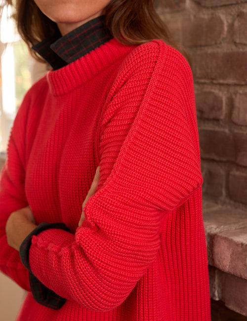 A close-up of a person wearing a bright red ribbed knit sweater over a green and red plaid collared shirt, standing in front of a brick wall., view 4
