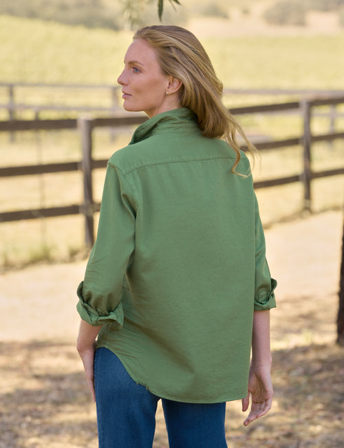 A woman wearing an olive green long-sleeve button-up shirt and blue jeans, photographed from the back in an outdoor rural setting with a wooden fence and dry grass., alternate view