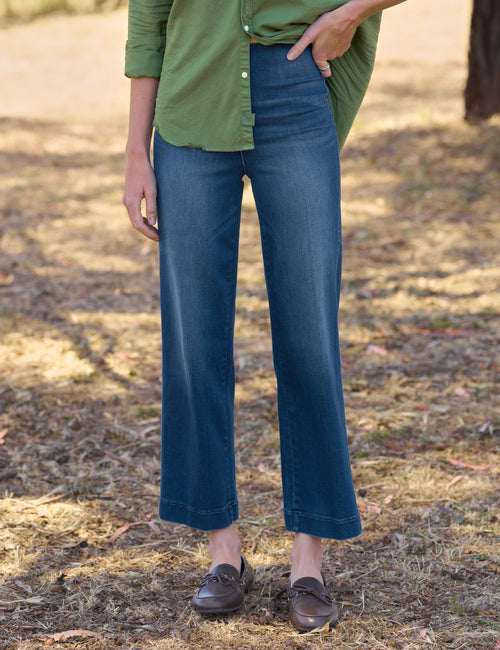 A person outdoors wearing dark blue cropped wide-leg jeans, a green button-up shirt, and brown leather loafers on a dry grassy path., front view