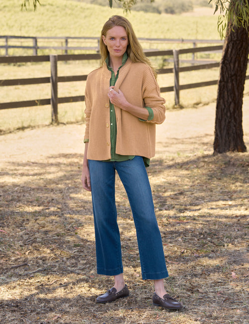 A woman stands outdoors in a sunlit field wearing a tan ribbed knit cardigan over a green button-up shirt, blue cropped jeans, and dark brown loafers., view 4