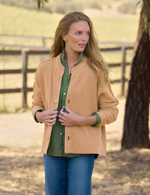A woman wearing a tan ribbed knit cardigan over an olive green button-up shirt and blue jeans, standing outdoors in front of a wooden fence and tree., front view